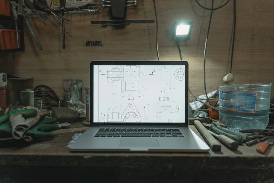 A laptop displaying blueprints on a cluttered workbench in a workshop setting.